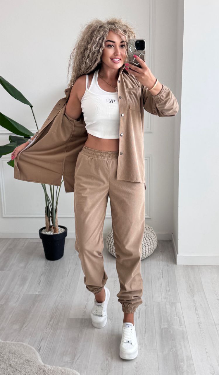 Woman taking a mirror selfie wearing a beige tracksuit and white sneakers in a room with a plant.