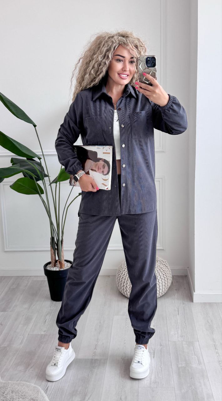 Woman taking a mirror selfie wearing a dark gray outfit with white sneakers in a room with a plant and decorative items.