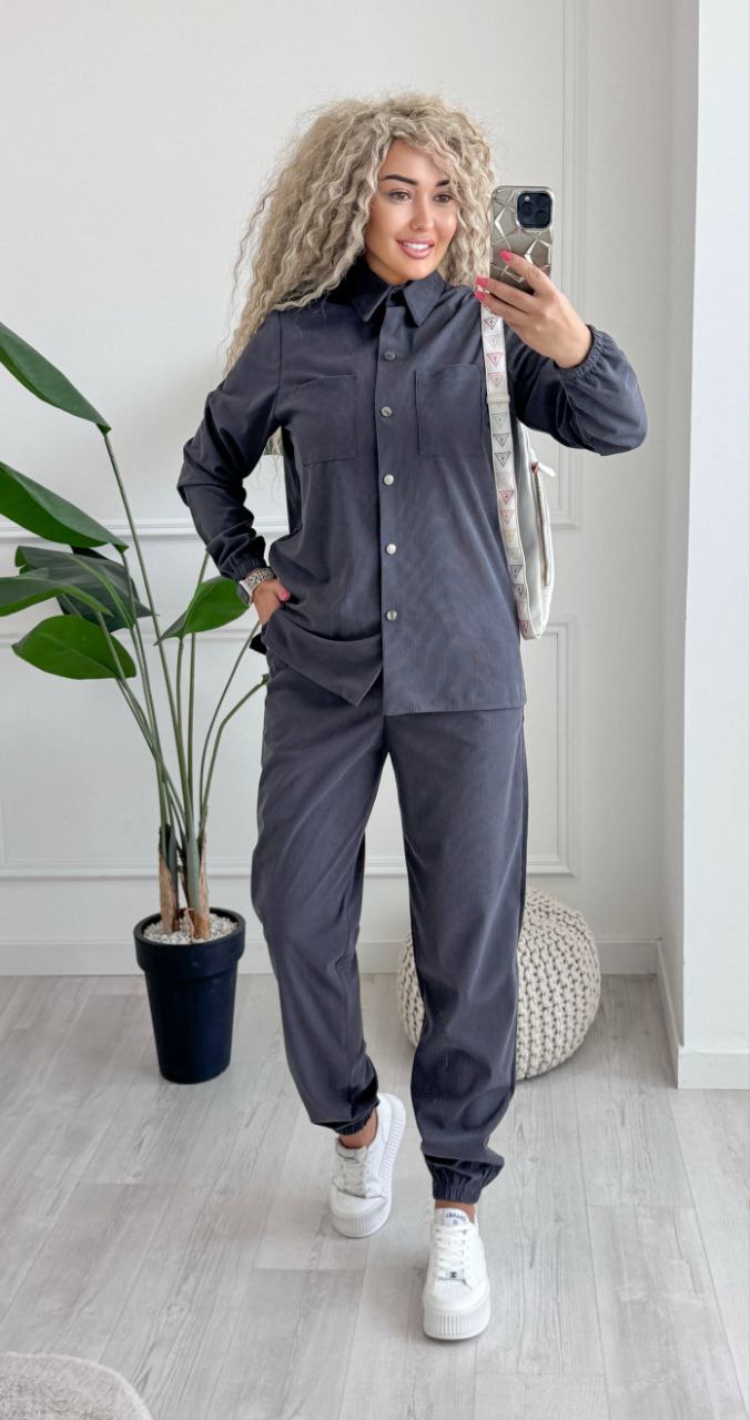 Person taking a mirror selfie wearing a dark gray outfit with white sneakers in a room with a plant and rug.