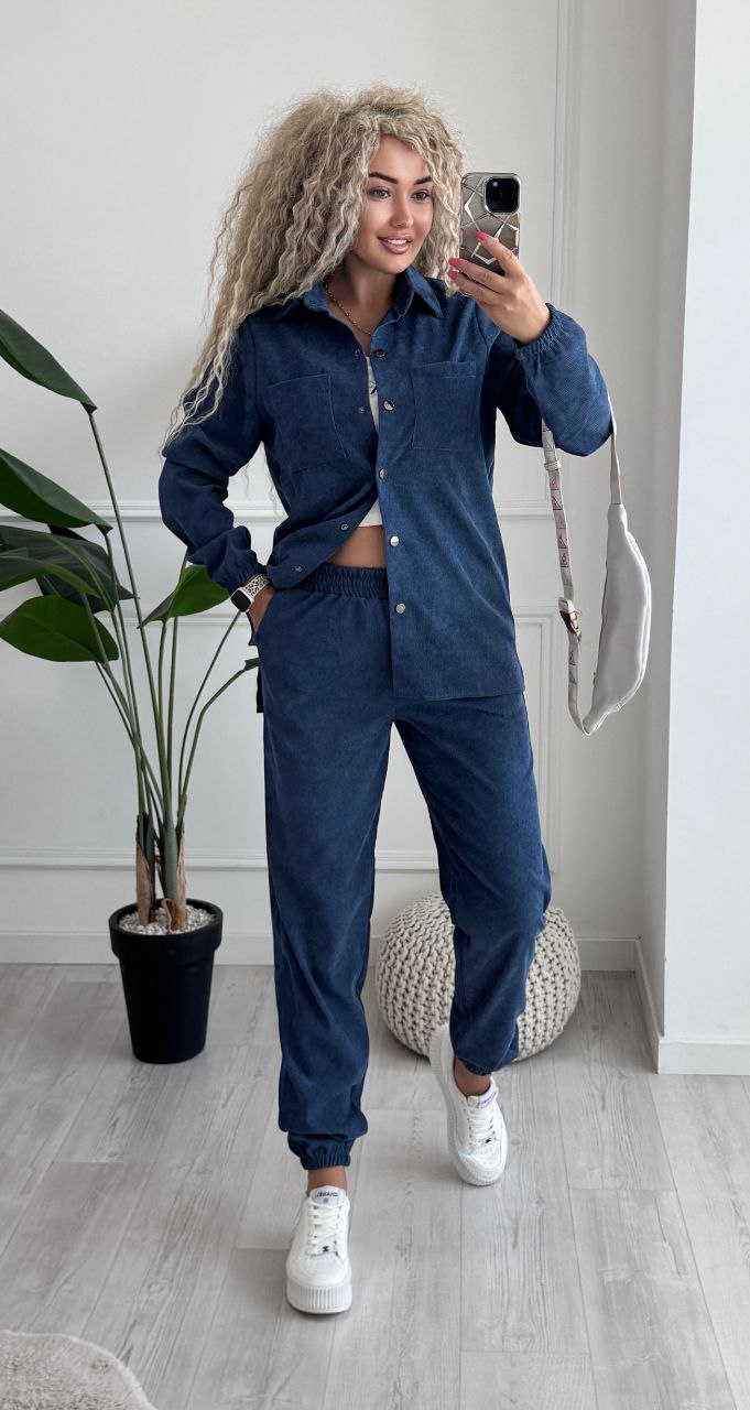 Person wearing a blue tracksuit taking a mirror selfie in a room with a plant and rug.