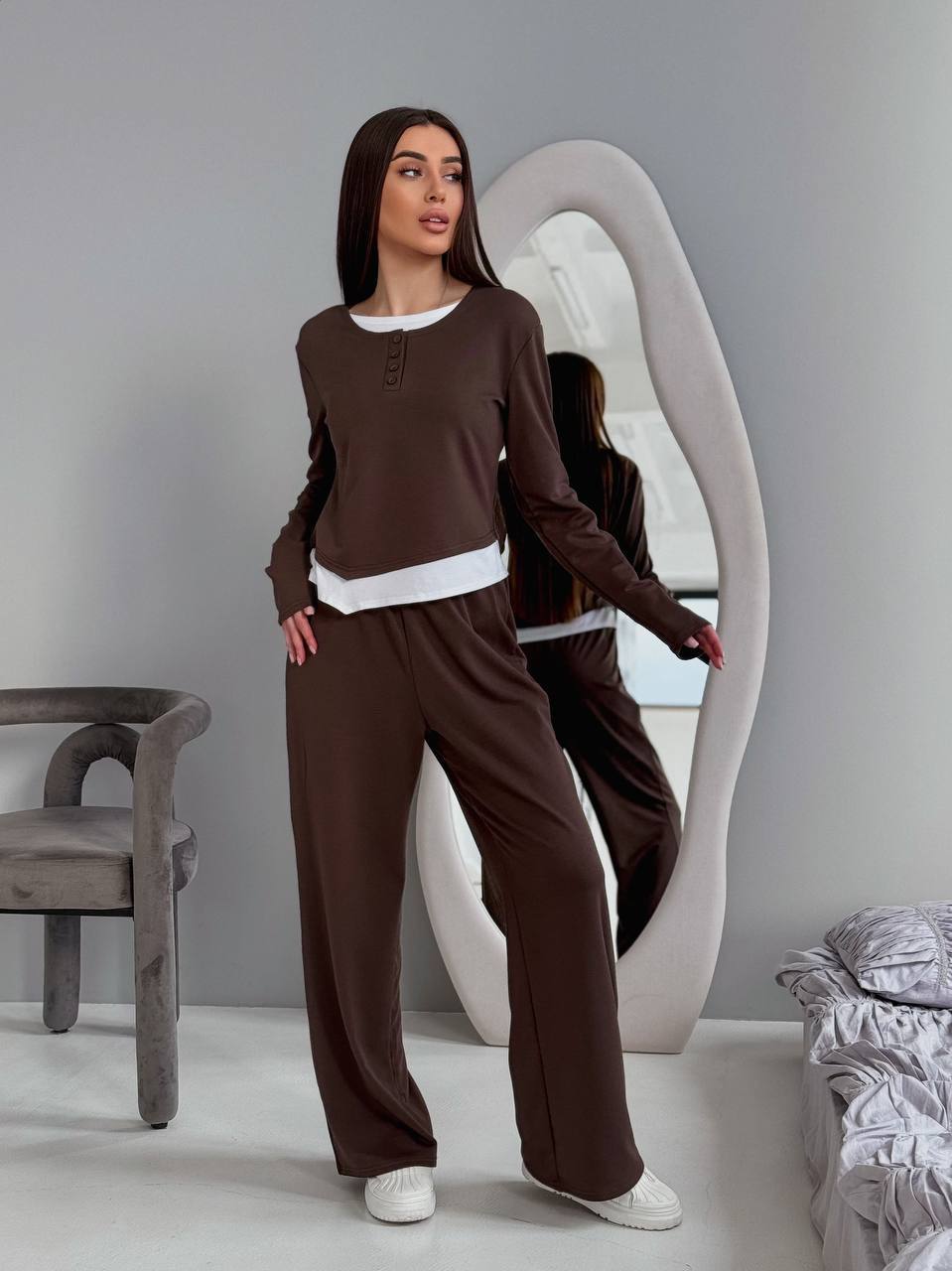 Woman in brown loungewear standing in a room with a mirror and chair.
