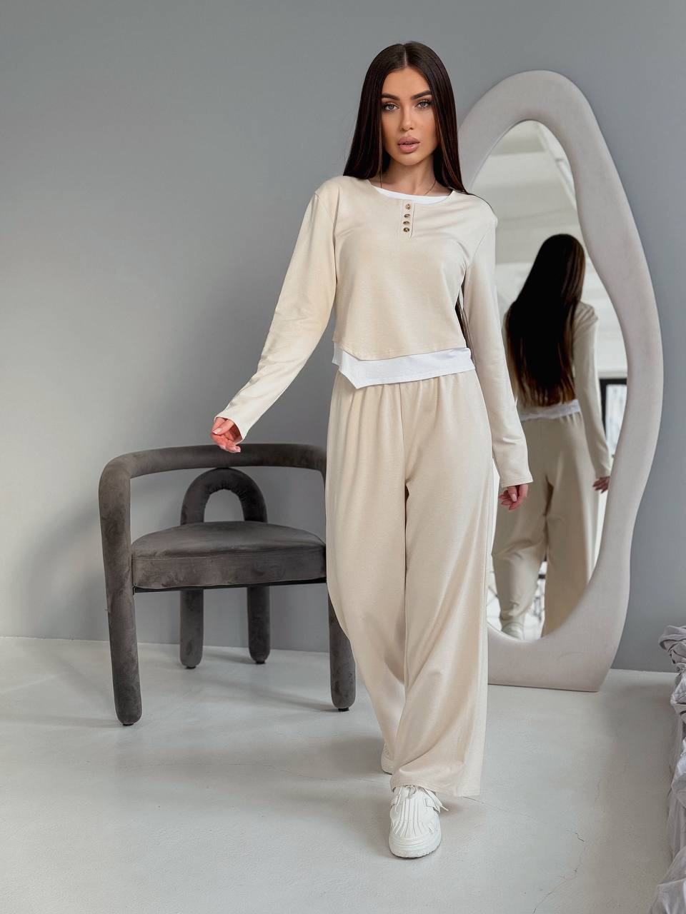 Woman in a beige outfit standing in a minimalistic room with a chair and mirror.