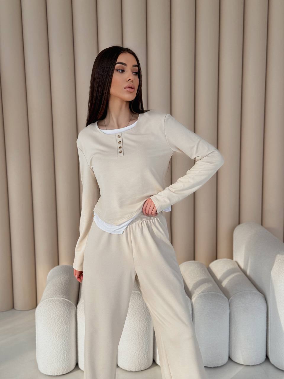 Woman wearing a beige long-sleeve top and pants in a neutral-colored room.