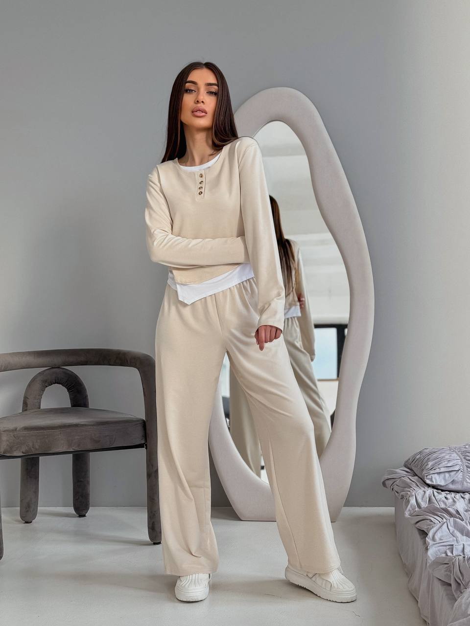 Woman wearing a beige outfit standing in a minimalistic room with a mirror and chair.