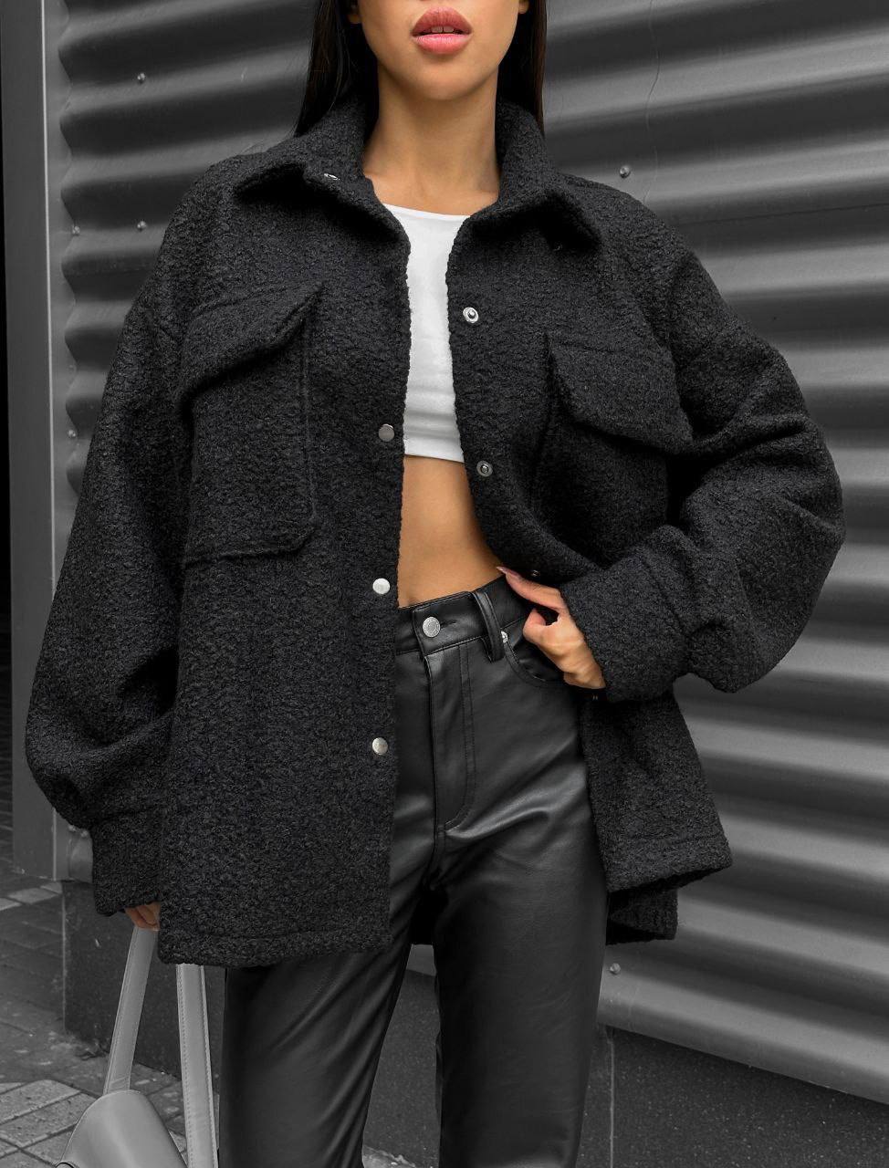Person wearing a dark gray coat over a white top and black pants against a corrugated metal background.