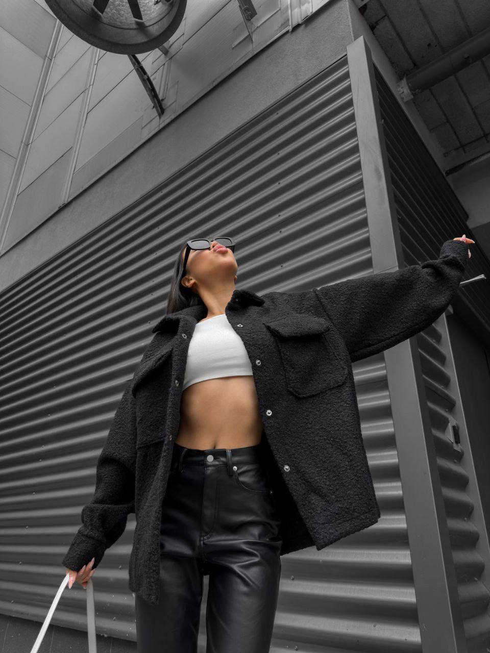 Person wearing a black jacket and leather pants standing against a gray building.