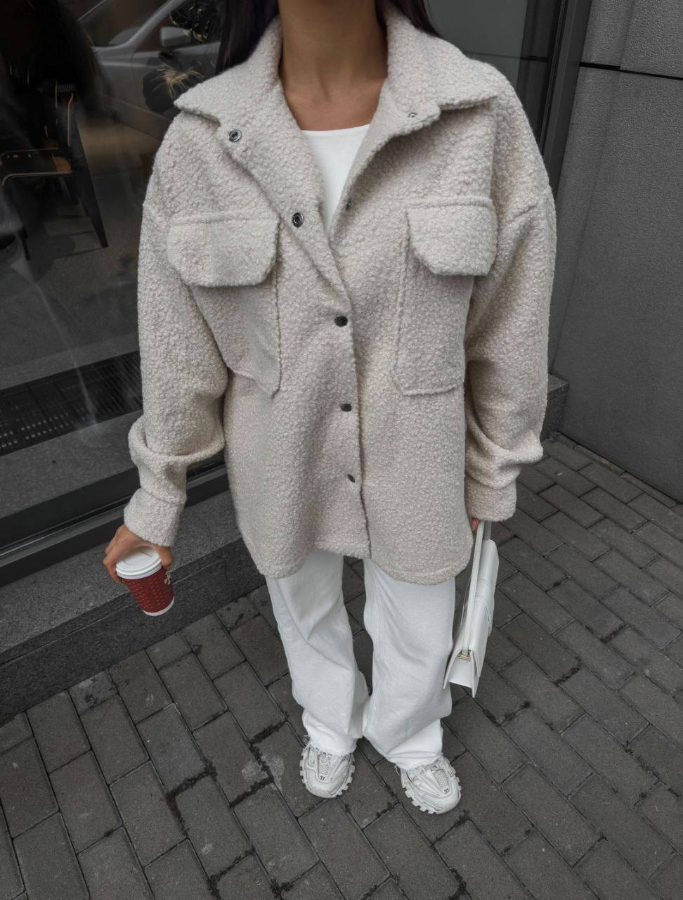 Person wearing a beige sherpa coat with a white shirt and pants, holding a red cup and a white bag.