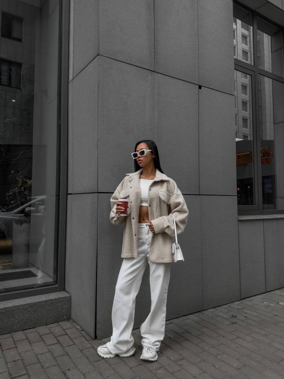 Woman in a beige coat and white pants standing against a gray building