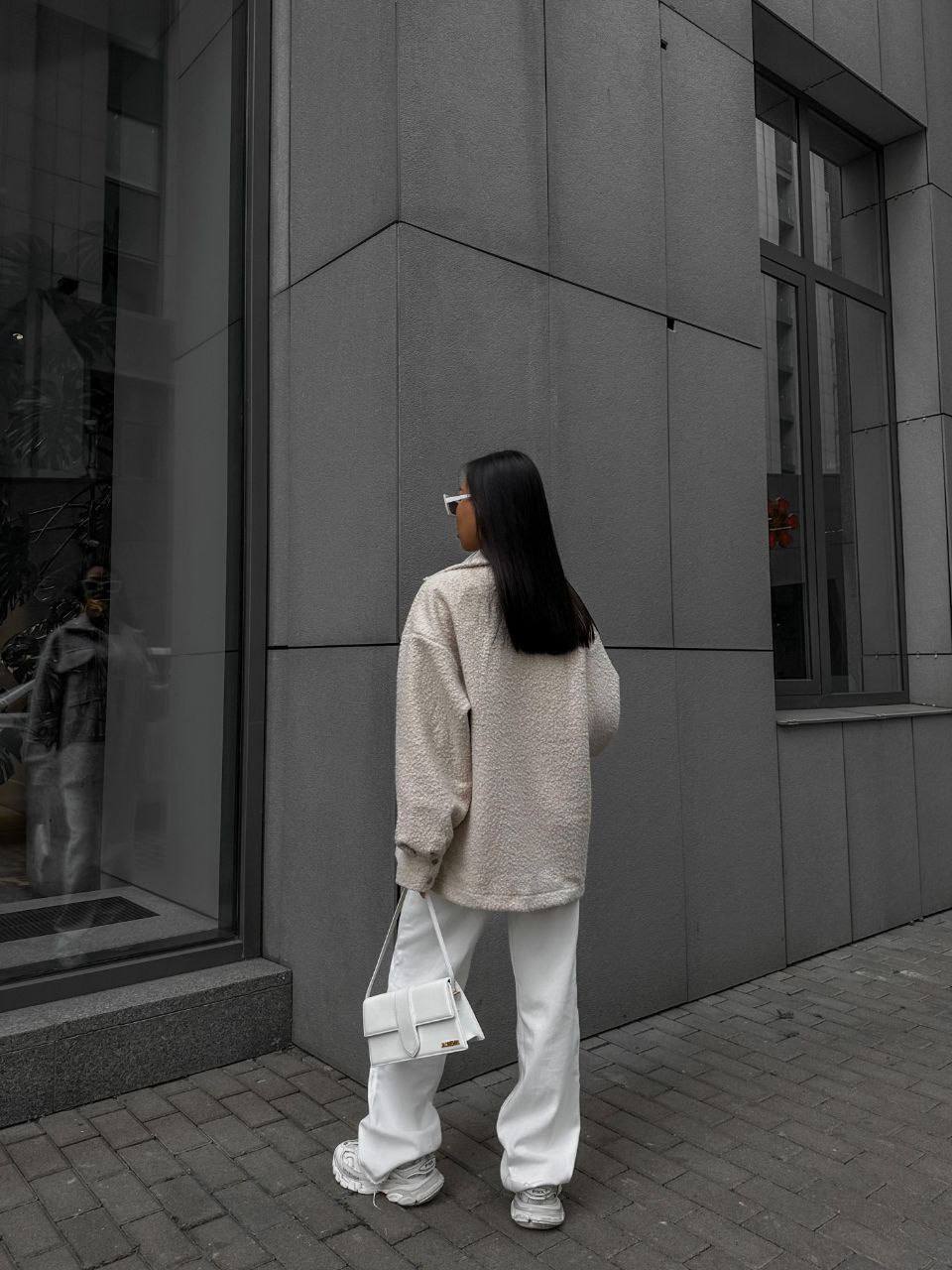 Person wearing a beige sweater and white pants standing in front of a gray building.