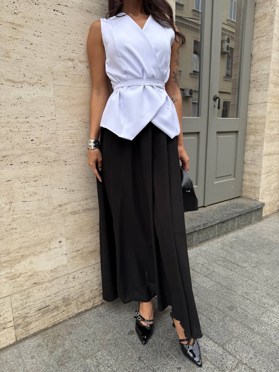 Woman wearing a white sleeveless top and black wide-leg pants against a beige wall.