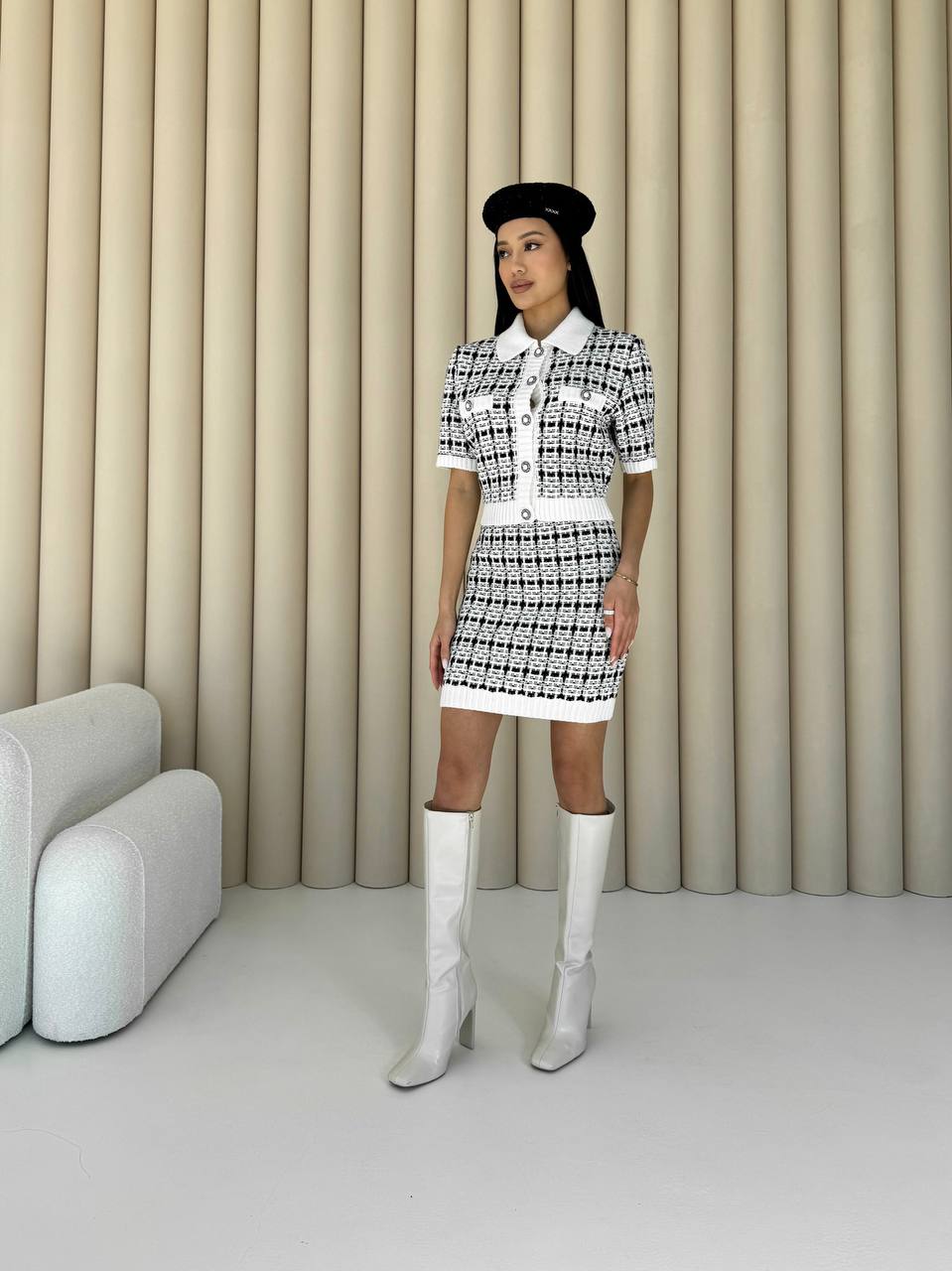 Woman in a black and white checkered dress with a beret and white boots standing against a beige curtain.