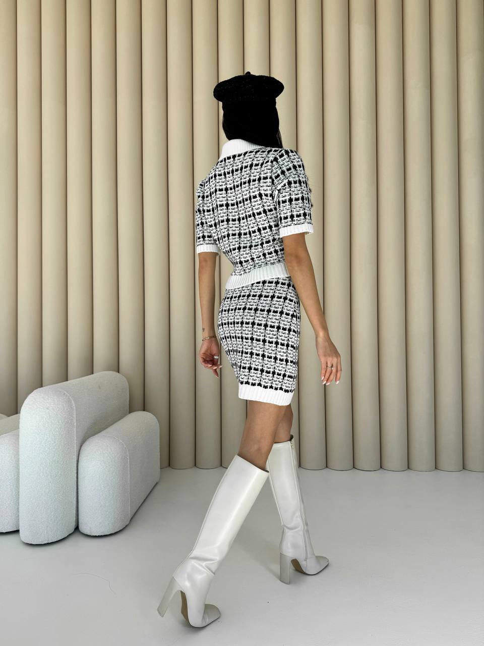 Person wearing a black and white patterned outfit with knee-high boots in a minimalistic room.