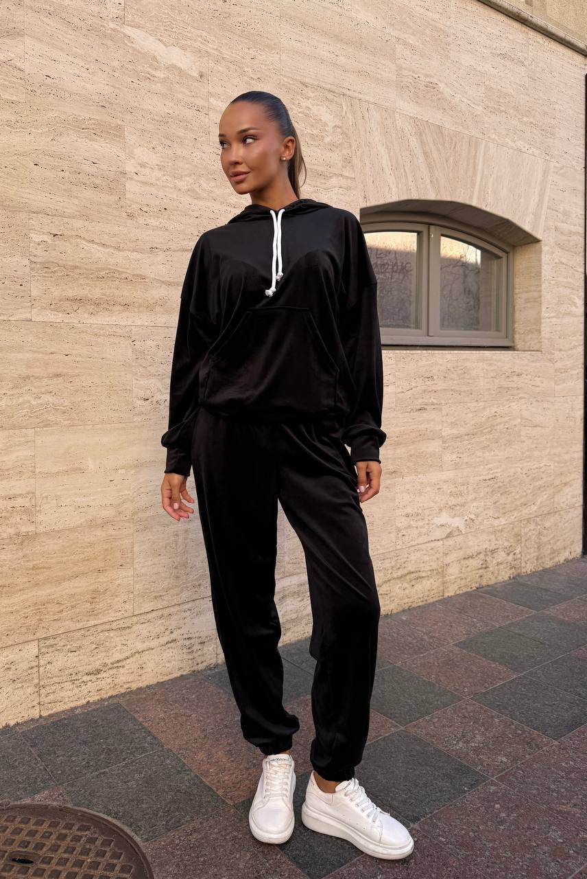 Person wearing a black tracksuit with white sneakers standing against a beige wall.