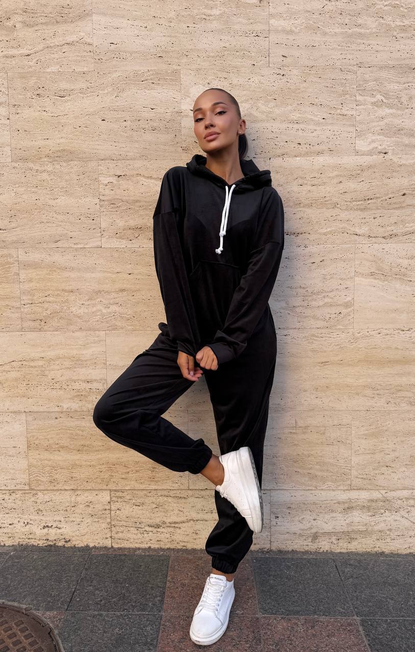 Person wearing a black tracksuit with white sneakers against a beige wall.
