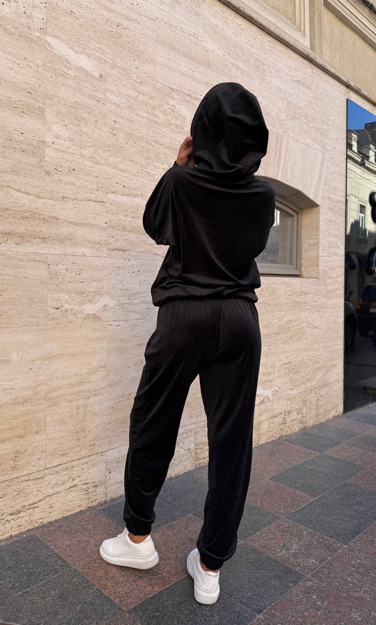 Person wearing a black hoodie and pants standing against a beige wall.