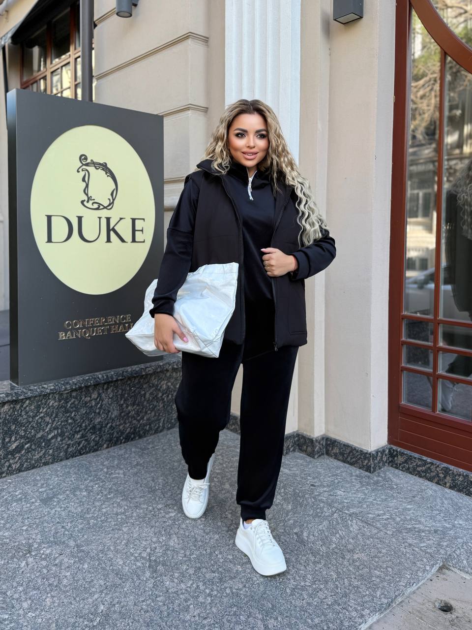 Woman in black outfit holding a white bag in front of a building with 'DUKE' signage.