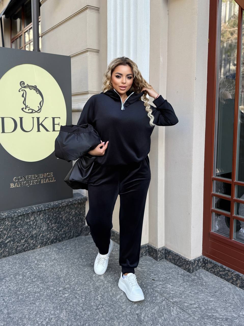 Woman in black outfit holding a black bag in front of a building with 'DUKE' signage.
