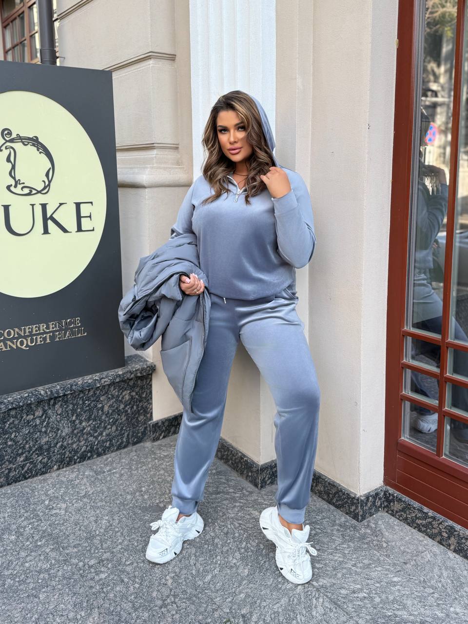Woman in a blue tracksuit standing outside a building with a sign that reads 'DUKE'.
