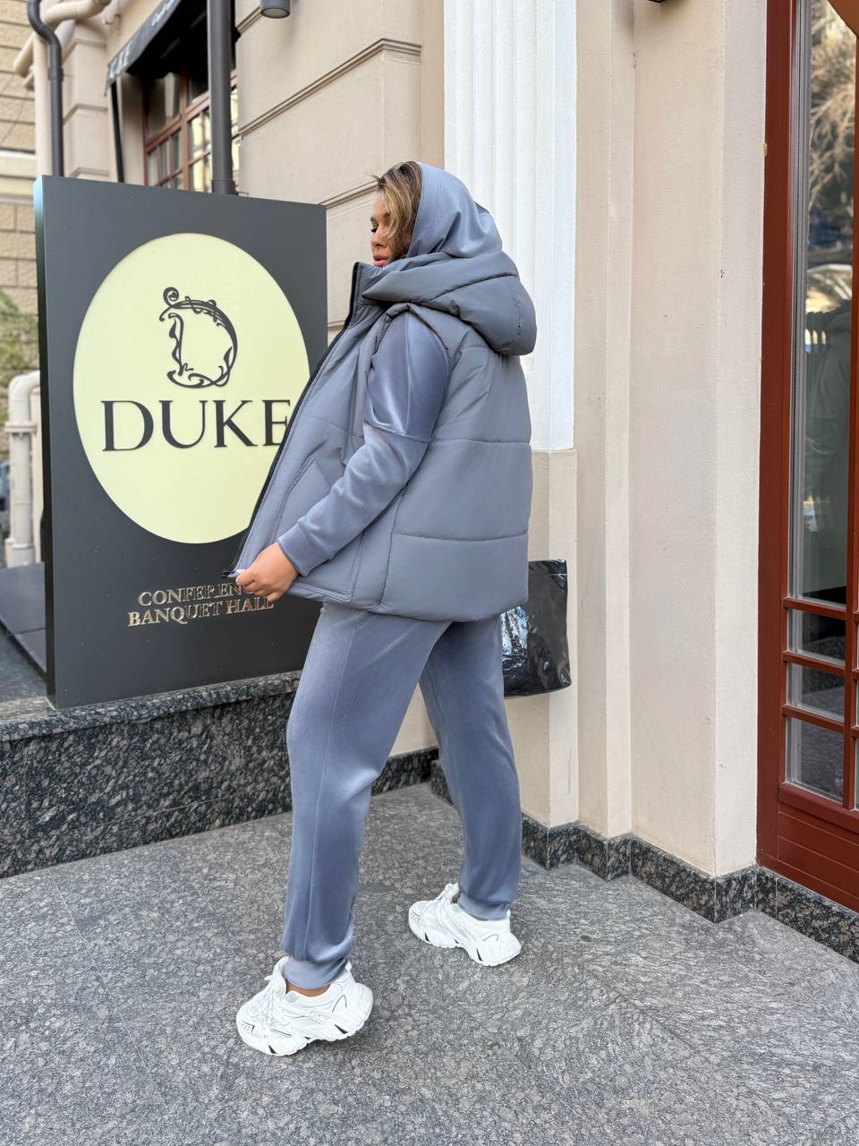 Person wearing a blue puffer jacket and pants in front of a building with a 'Duke' sign.