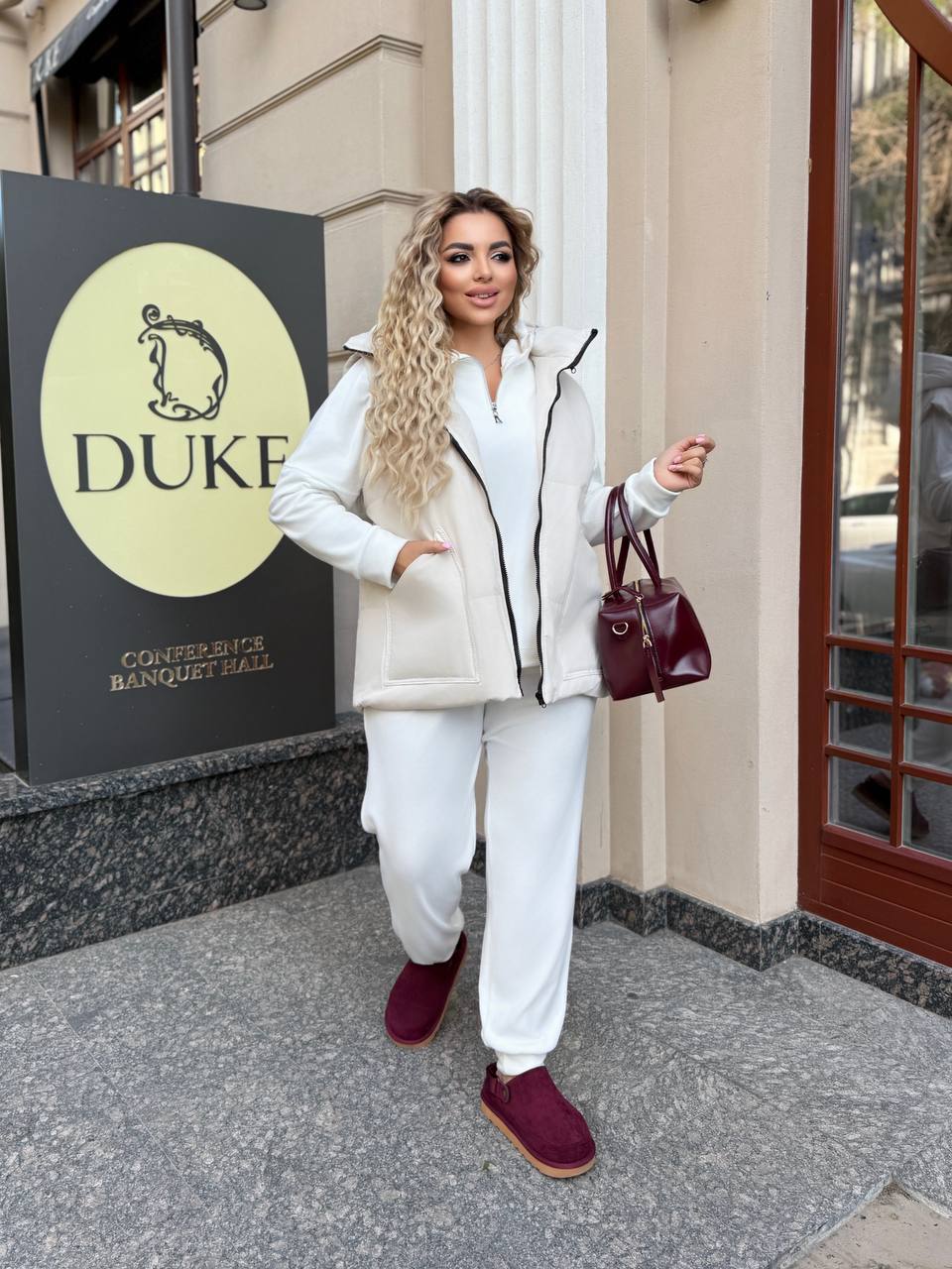 Woman in white outfit with burgundy shoes standing in front of a building with 'DUKE' sign.