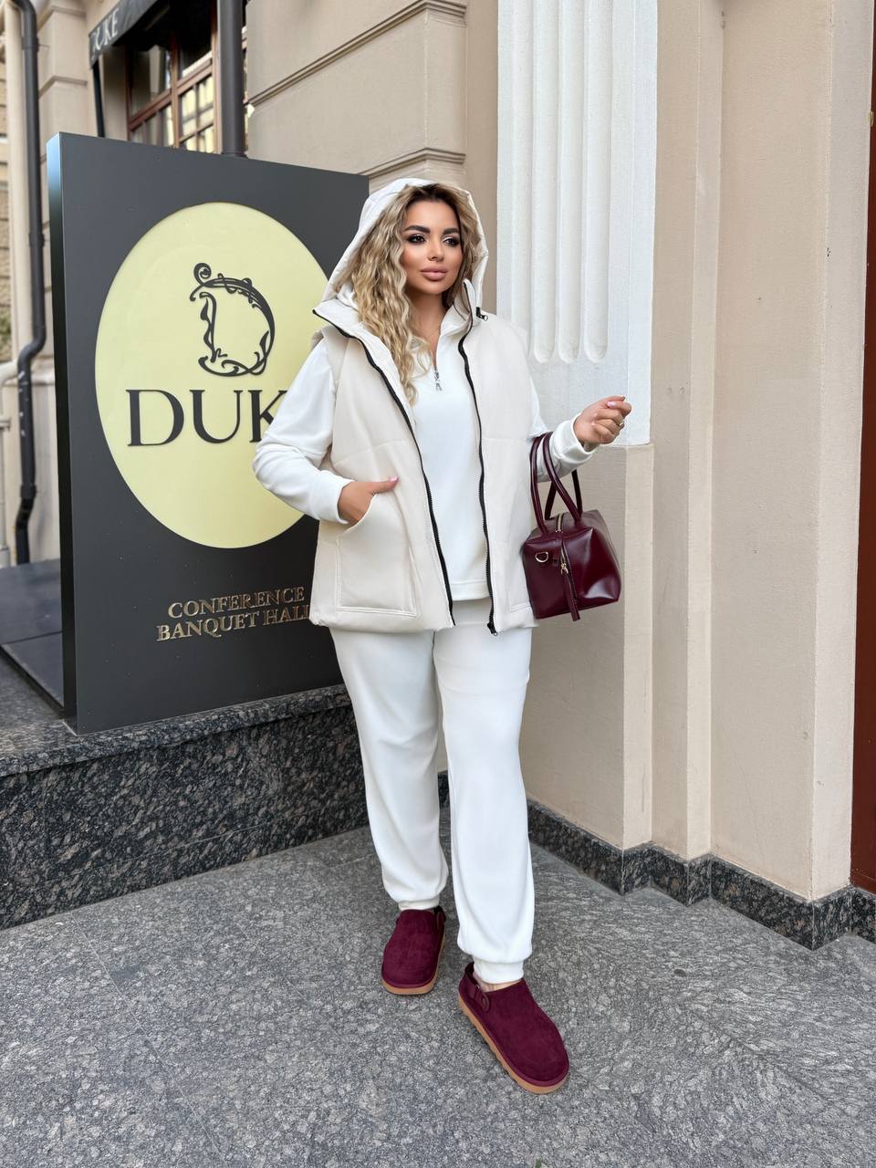 Woman in a white outfit with maroon shoes standing in front of a building with a 'DUK' sign.