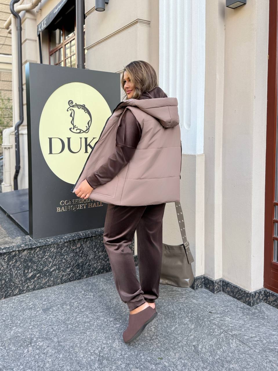Woman in a brown tracksuit standing in front of a building with a sign that reads 'DUK'.