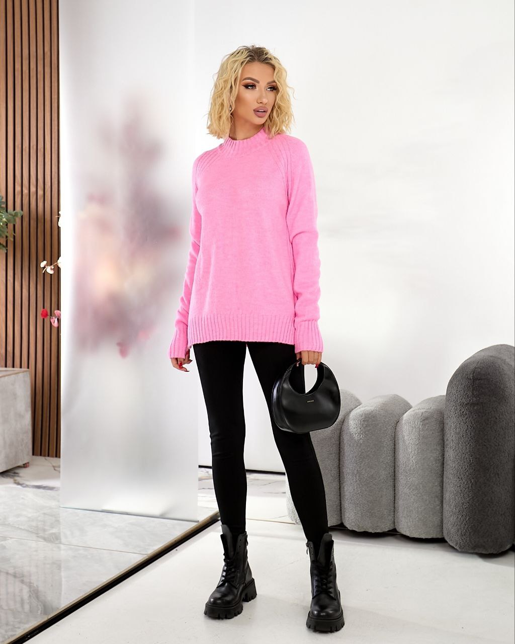 Woman wearing a pink sweater and black pants in a modern room.