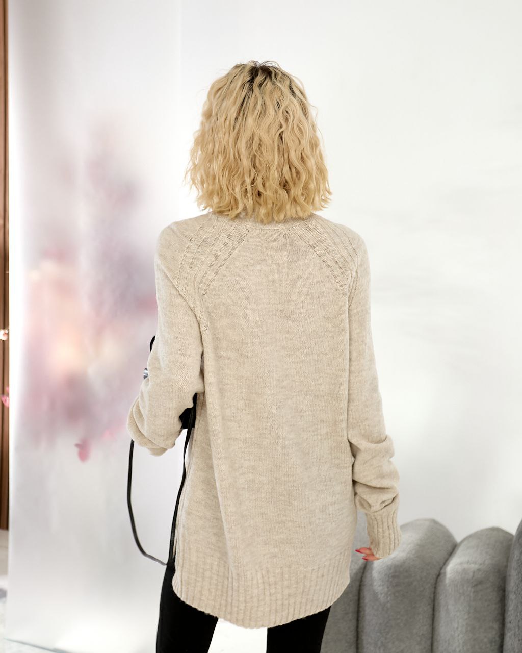 Person wearing a beige cardigan in a room with a white wall and gray sofa.