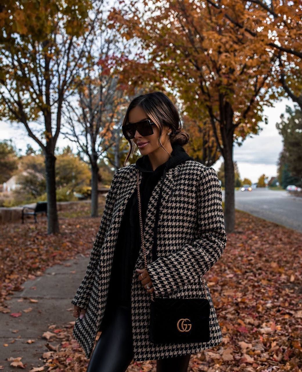 Woman in a houndstooth coat with a black handbag walking on a leaf-covered sidewalk.