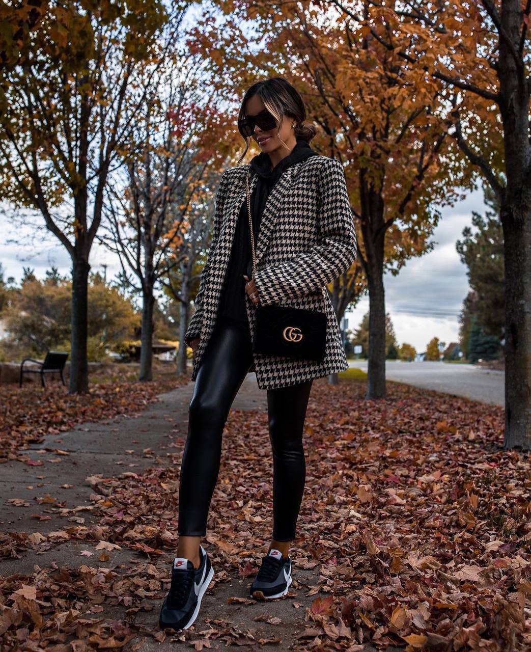 Person wearing a houndstooth coat and black leggings standing on a leaf-covered path with trees in the background.