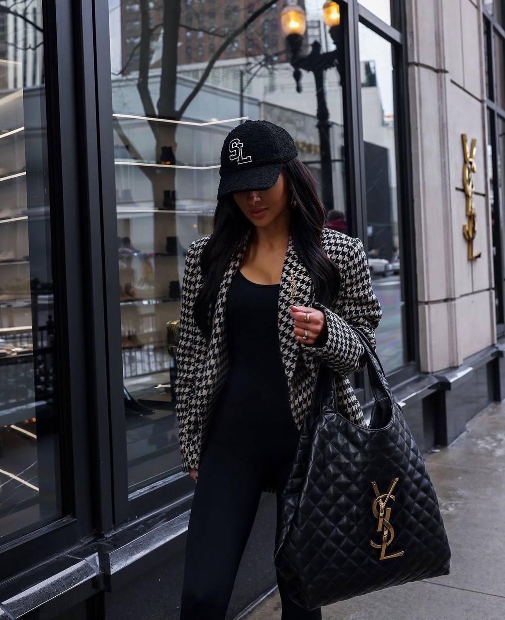 Woman in a houndstooth blazer and black cap holding a YSL bag on a city street.