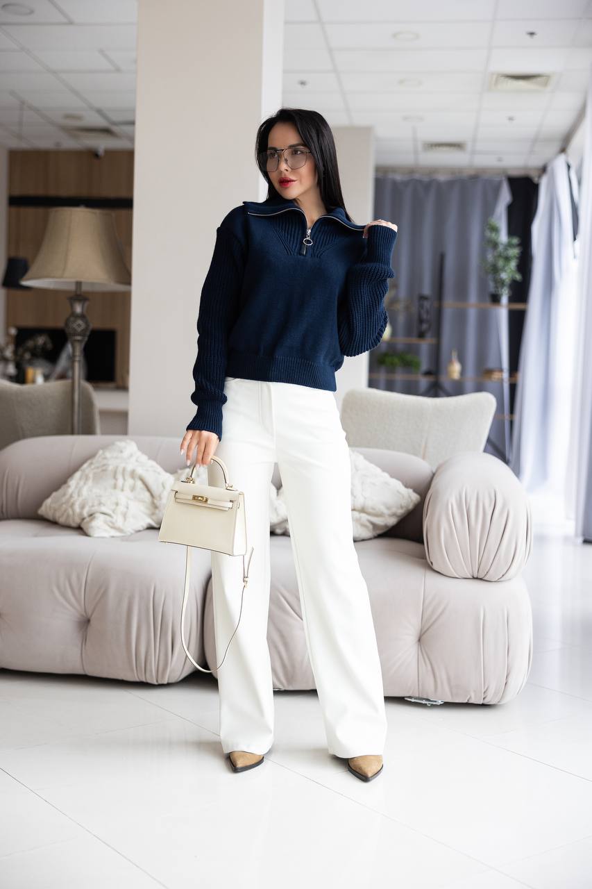 Woman in navy sweater and white pants standing in a modern living room.