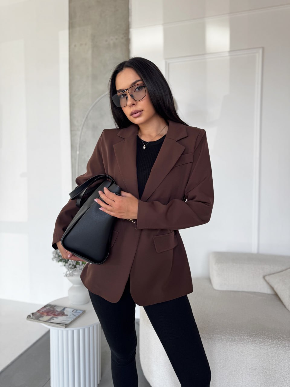 Woman in a brown blazer holding a black handbag in a modern interior setting