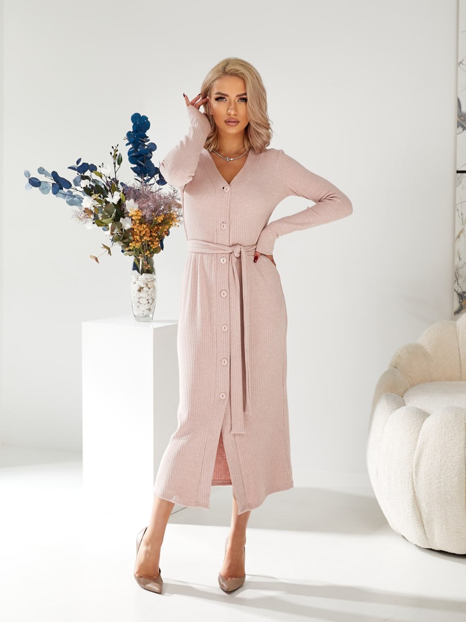 Woman wearing a pink dress standing in a minimalistic room with a vase of flowers and a white pumpkin.