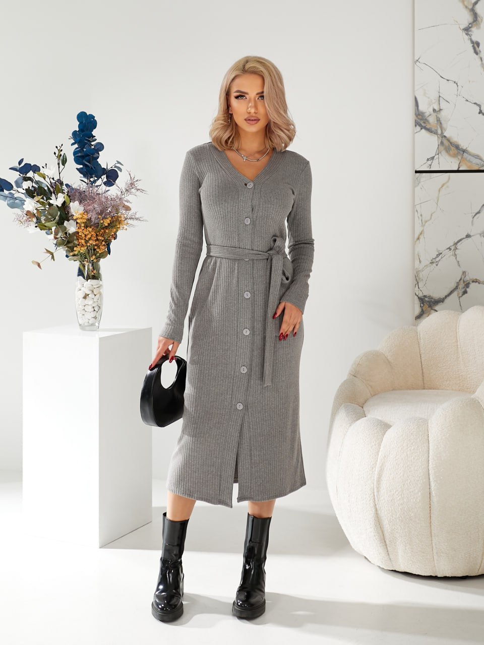 Woman wearing a gray dress standing in a modern interior setting with a white armchair and decorative flowers.