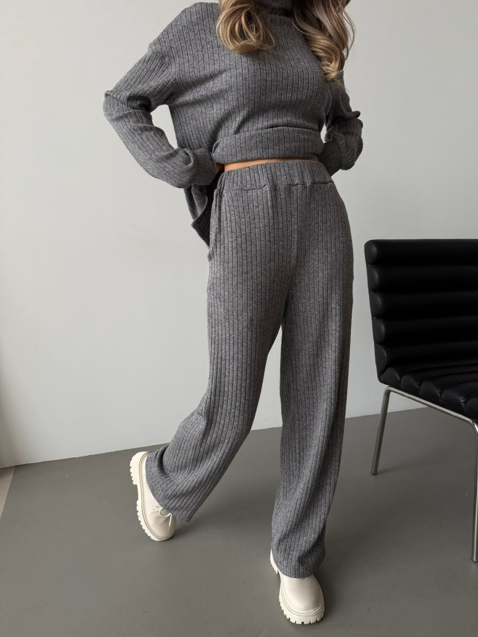 Person wearing a gray knit outfit standing in a minimalistic room with a chair.