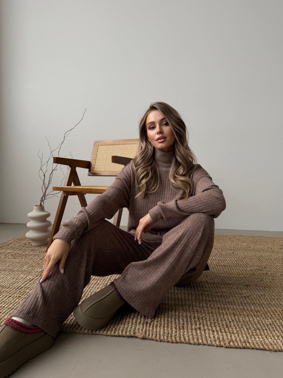 Woman sitting on a rug wearing a brown sweater and pants in a minimalistic room.