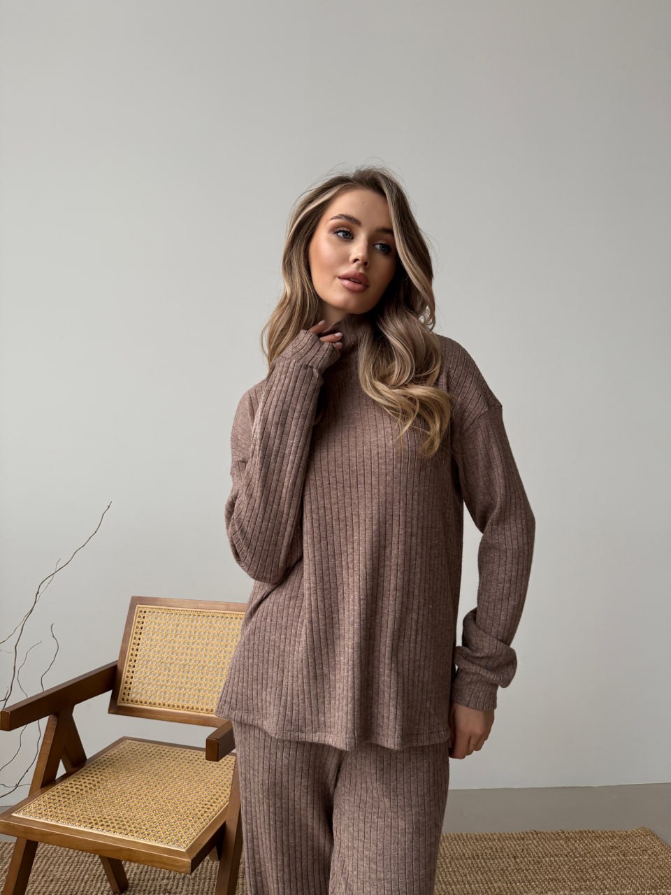 Woman wearing a brown ribbed sweater and pants standing in a room with a chair and decorative branch.