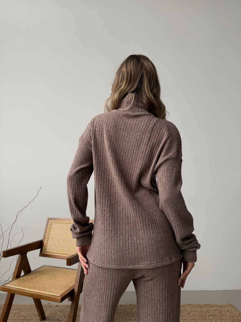 Person wearing a brown ribbed knit outfit standing in a minimalistic room with a chair and plant.