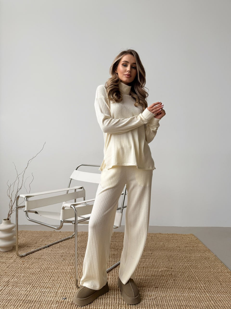 Woman wearing a cream-colored outfit standing in a minimalistic room with a chair and vase.
