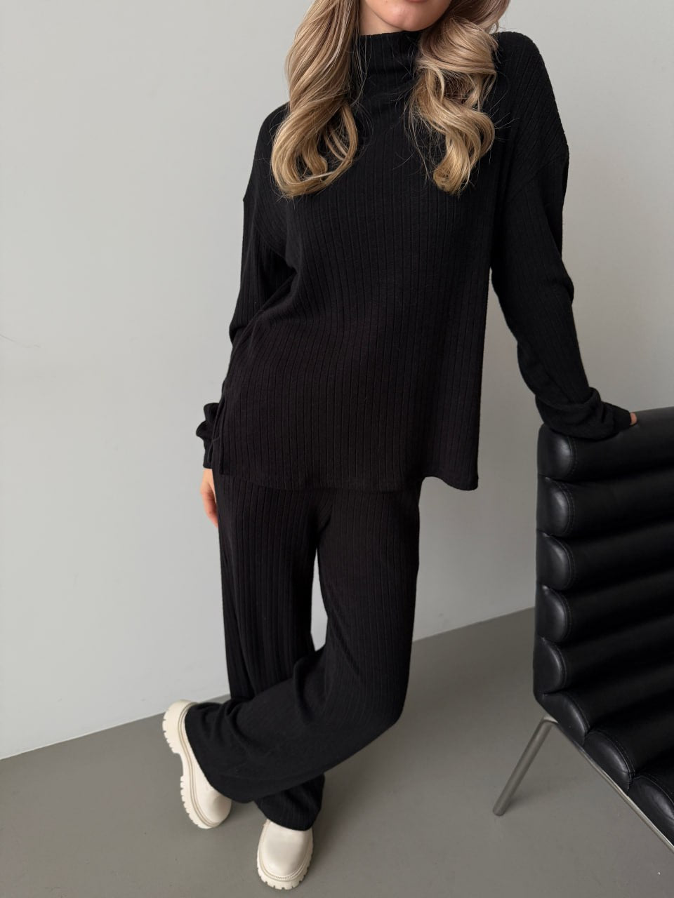 Person wearing a black ribbed sweater and pants with white shoes, standing against a gray wall.
