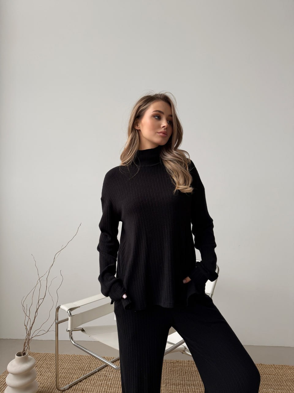 Woman wearing a black turtleneck sweater and pants in a minimalistic setting.