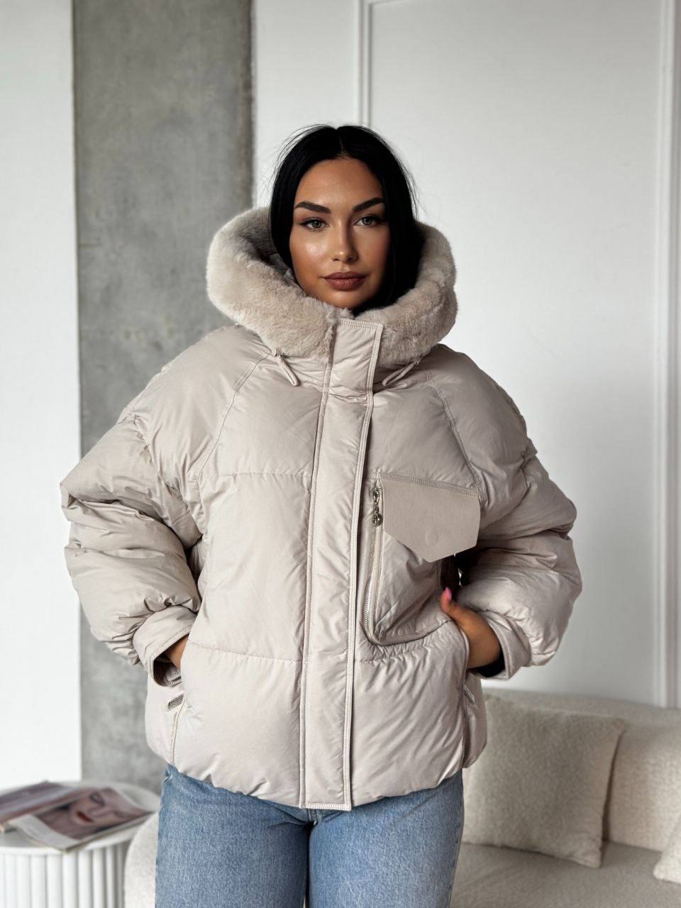 Woman wearing a beige puffer jacket with a fur-lined hood in a room.
