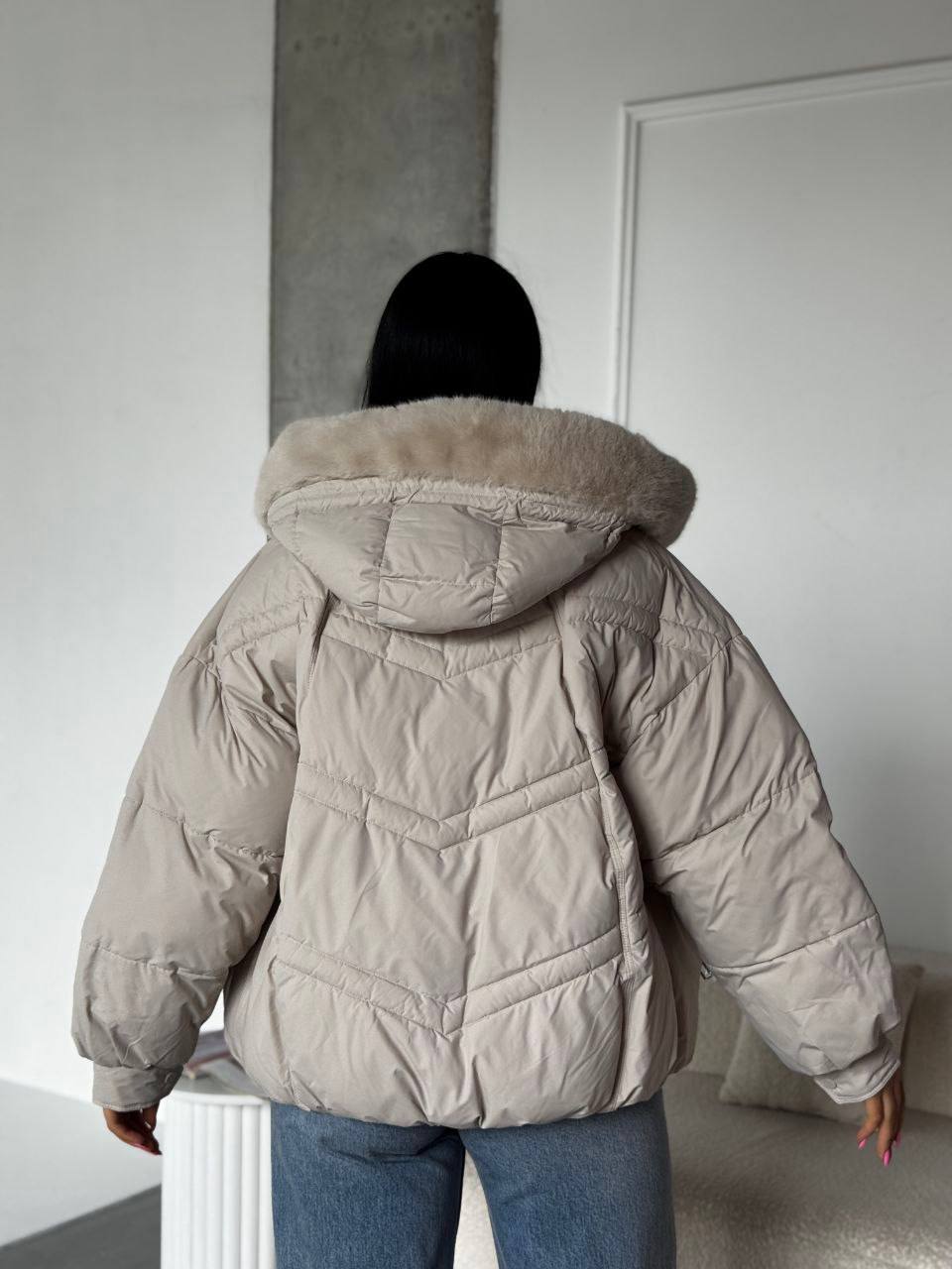 Person wearing a beige puffer jacket with a fur-lined hood in a minimal indoor setting.