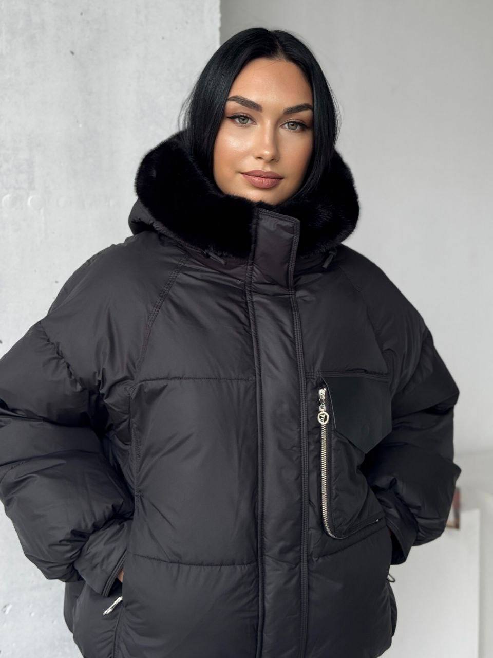 Woman wearing a black puffer jacket with fur hood against a white wall