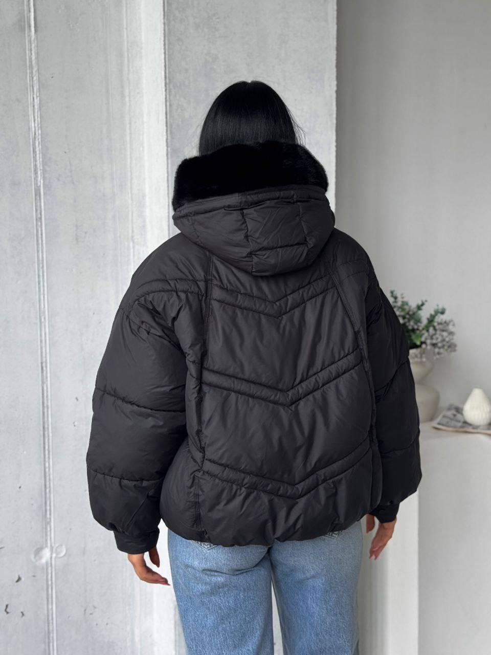 Person wearing a black puffer jacket with a fur-lined hood against a light gray wall.