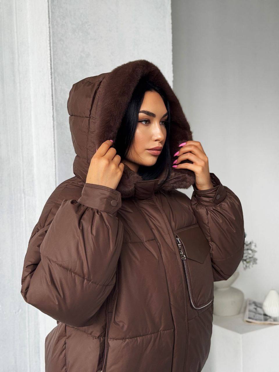 Woman wearing a brown puffer coat with a hood indoors.