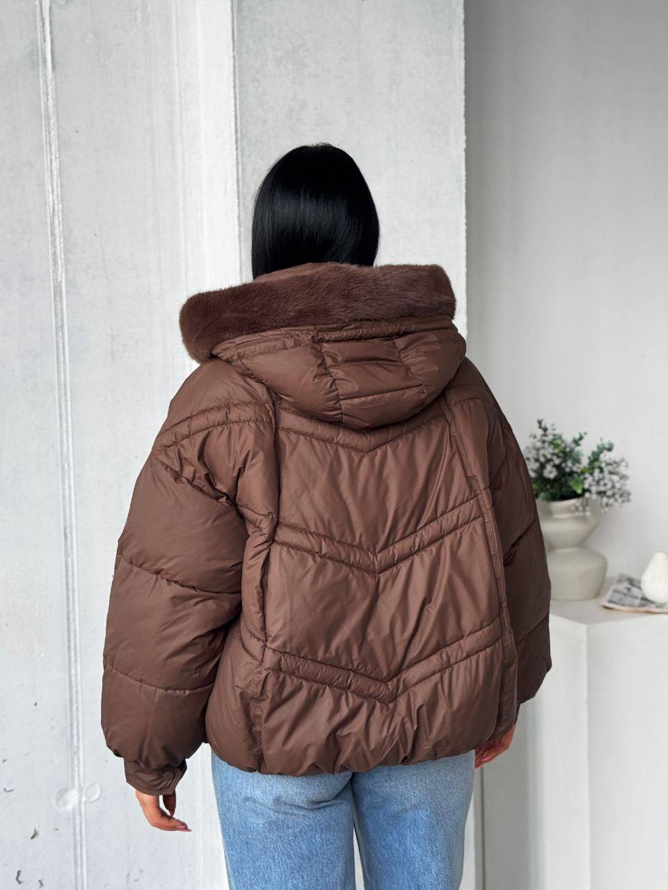 Person wearing a brown puffer jacket with fur hood indoors.