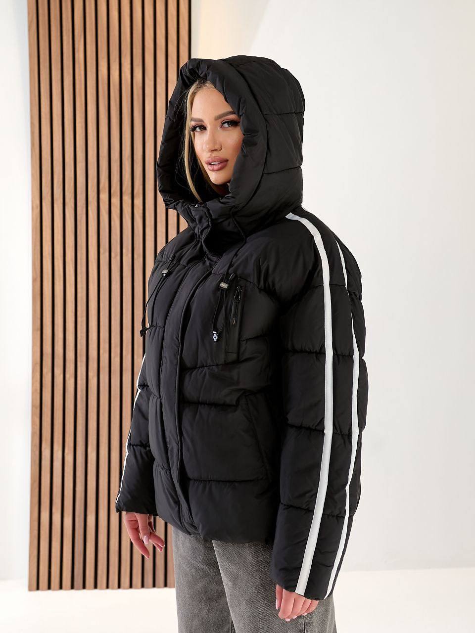 Person wearing a black puffer jacket with a hood and white stripes on a plain background