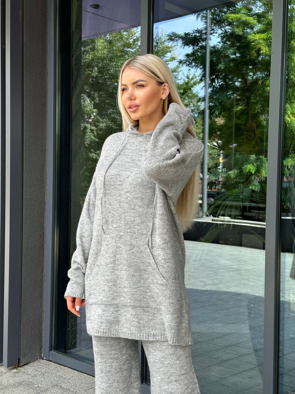 Woman wearing a gray hoodie and pants standing outside a building with reflective glass doors.